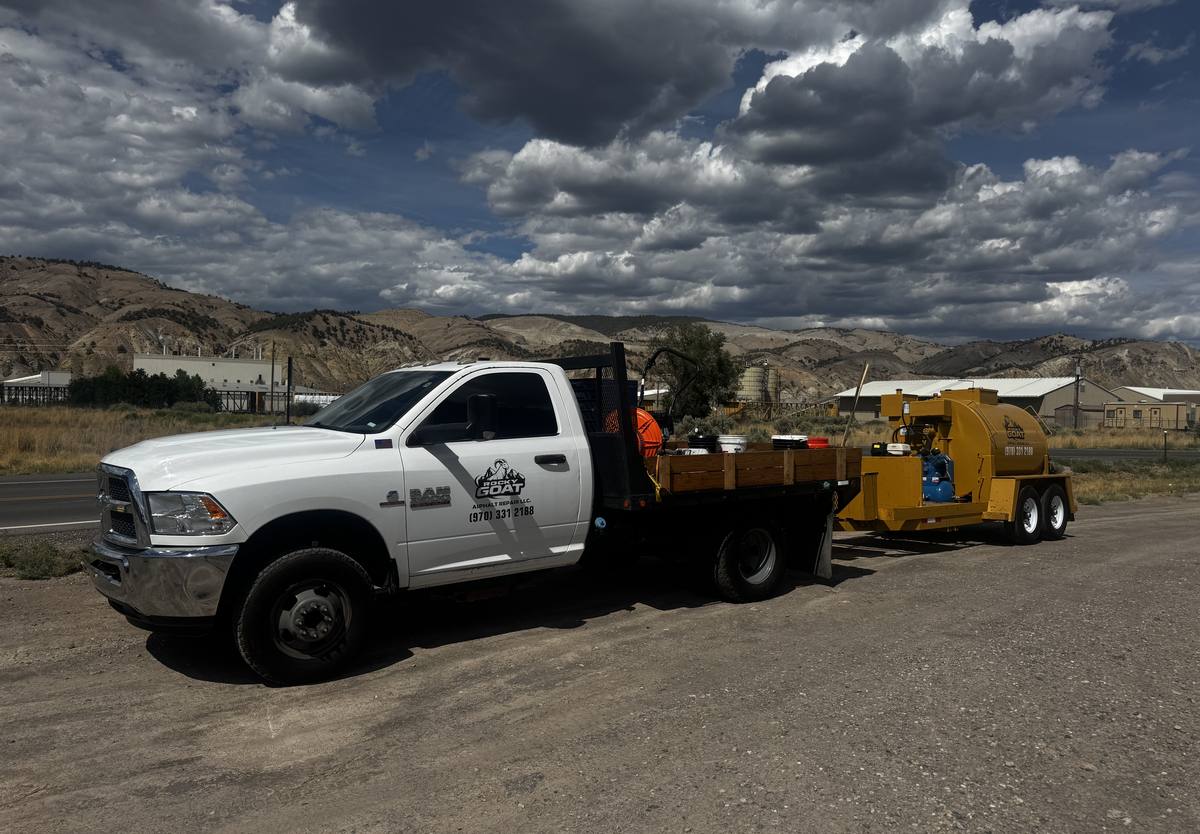 The Rocky Goat flatbed truck and a yellow sealcoat tank trailer parked on a gravel shoulder, both marked with the company logo and phone number